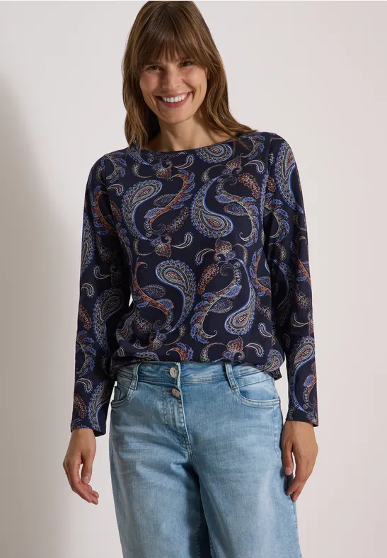 Lächelnde Frau mit blauem Paisley-Oberteil und hellblauen Jeans vor neutralem Hintergrund