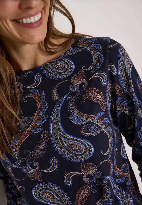 Nahaufnahme einer Frau im dunkelblauen Shirt mit buntem Paisley-Muster und leichtem Lächeln.