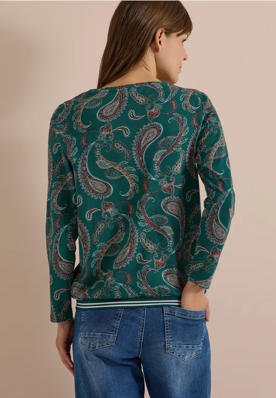 Frau von hinten in grünem Pullover mit Paisley-Muster und blauer Jeans.