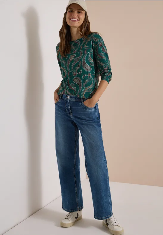 Eine junge Frau trägt Jeans, weiße Sneaker und eine grüne gemusterte Bluse, sie lächelt in neutralem Umfeld.