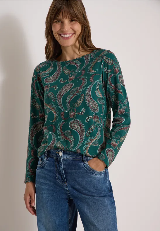 Frau in grünem Paisley-Oberteil und blauen Jeans, lächelt mit Händen in den Hosentaschen.