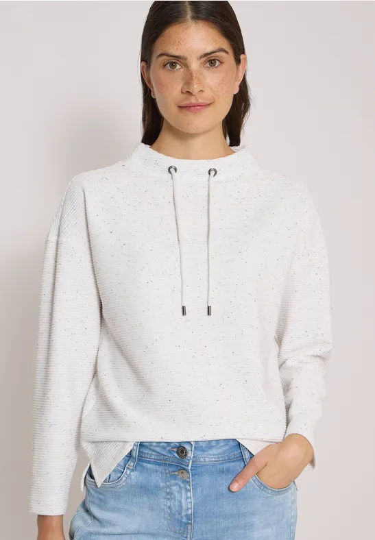 Junge Frau in hellem Kapuzenpullover mit Kordeln und Jeans, vor neutralem Hintergrund.