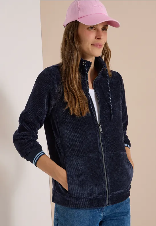 Frau in dunkelblauer Fleece-Jacke, weißem Shirt, Jeans und rosa Cap vor hellem Hintergrund.