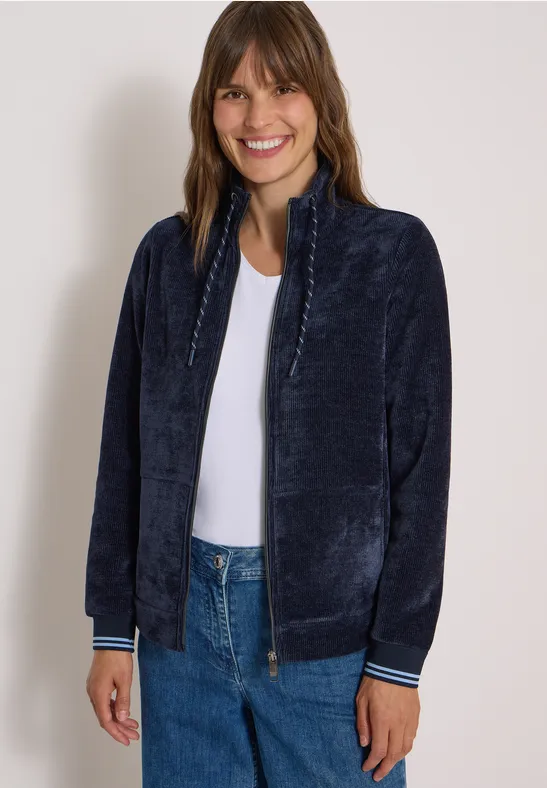Frau mit mittellangem braunem Haar trägt eine dunkelblaue Plüsch-Jacke, weiße Bluse und Jeans, lächelnd.