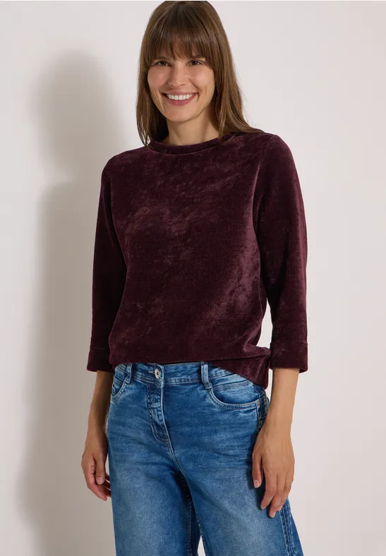 Frau in purpurnem Samtoberteil und blauer Jeans mit lächelnder Pose.