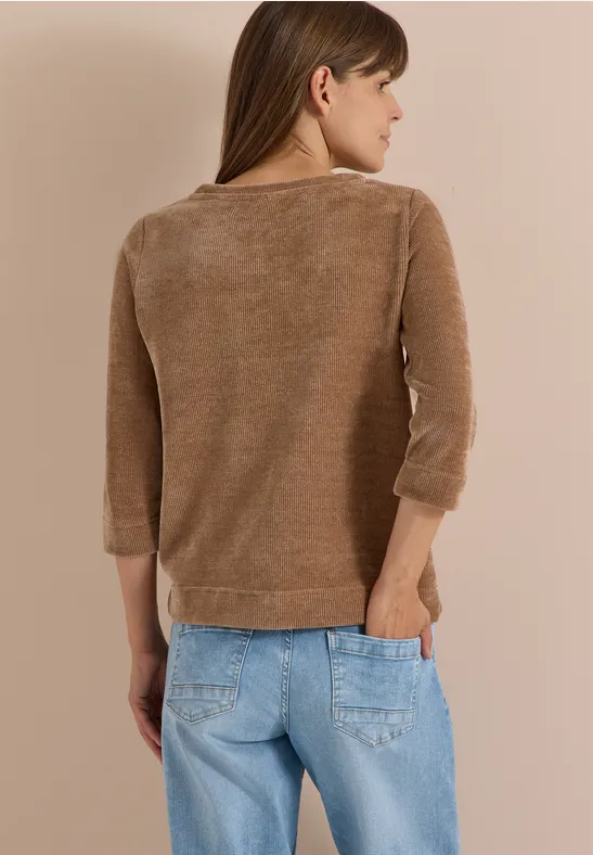 Vrouw van achteren in bruine trui en lichtblauwe jeans, zijwaarts kijkend naar rechts op een neutrale achtergrond.