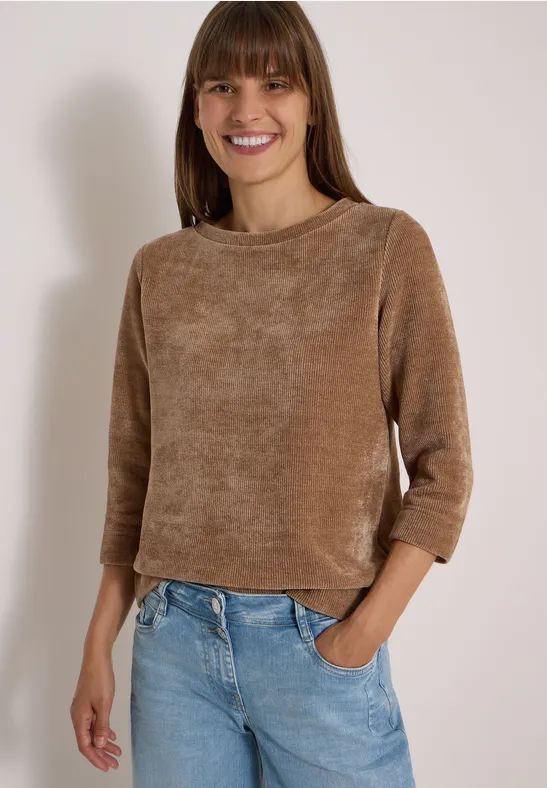 Een lachende vrouw in een bruine chenilletrui en lichtblauwe jeans, hand in zak, voor een lichte achtergrond.