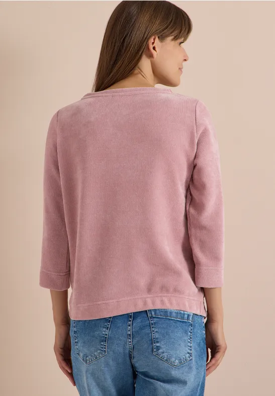 Person von hinten in rosafarbenem Kuschelpullover mit 3/4-Ärmeln und blauen Jeans vor beigem Hintergrund.