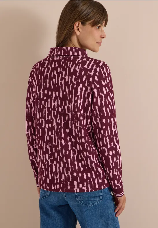 Frau mit braunen Haaren, in lila-pink gemustertem Langarmshirt und blauen Jeans, seitlich stehend