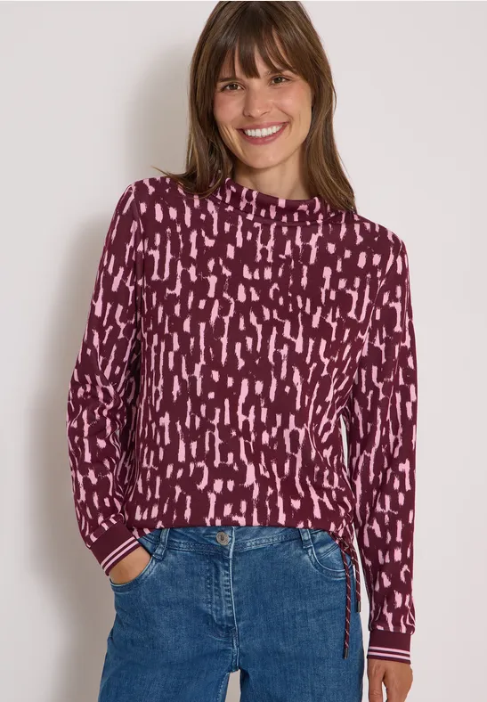 Eine lächelnde Frau in burgunderfarbenem Pullover mit abstraktem Muster und blauen Jeans.