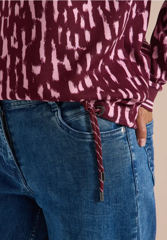 Person trägt lila gemustertes Oberteil mit Tunnelzug an der Seite und blaue Jeans.