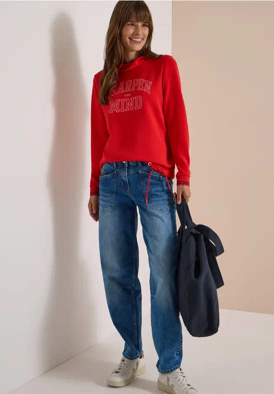 Model in rotem Sweatshirt mit Aufdruck, weiter Jeans und weißen Sneakern, hält eine dunkelblaue Jacke.