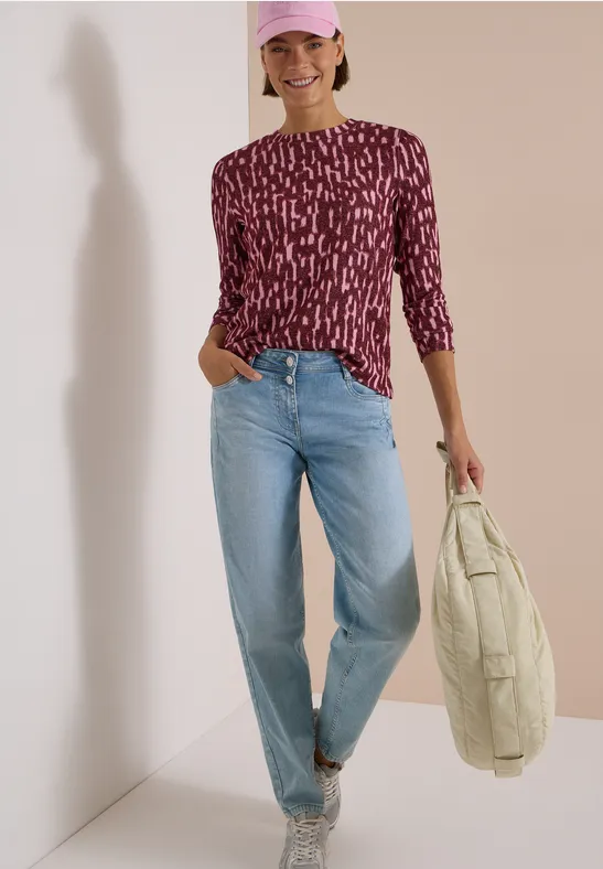 Frau mit lila gemustertem Shirt, hellen Jeans, Cap und gestepptem Beutel, lächelnd an einer Wand entlang.