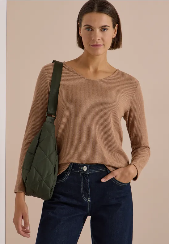 Frau mit beigem Pullover, blauen Jeans und grünem, gestepptem Schulterbeutel.