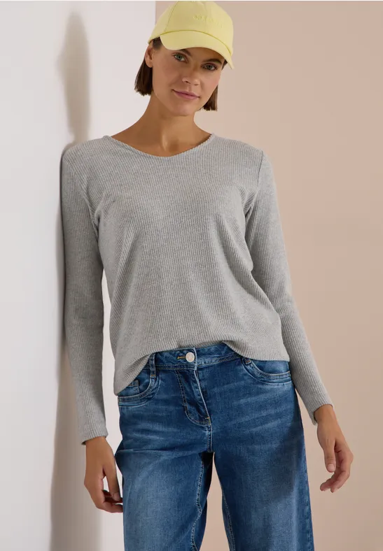 Frau mit grauem Langarmshirt, blauen Jeans und gelber Cap, stehend vor hellem Hintergrund.