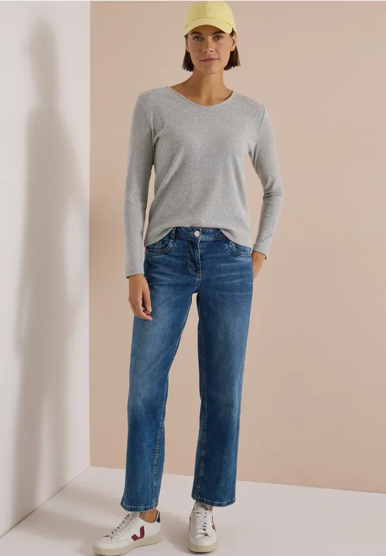 Frau in blauer Jeans, grauem Langarmshirt, weißen Sneakers und gelber Cap vor neutralem Hintergrund.