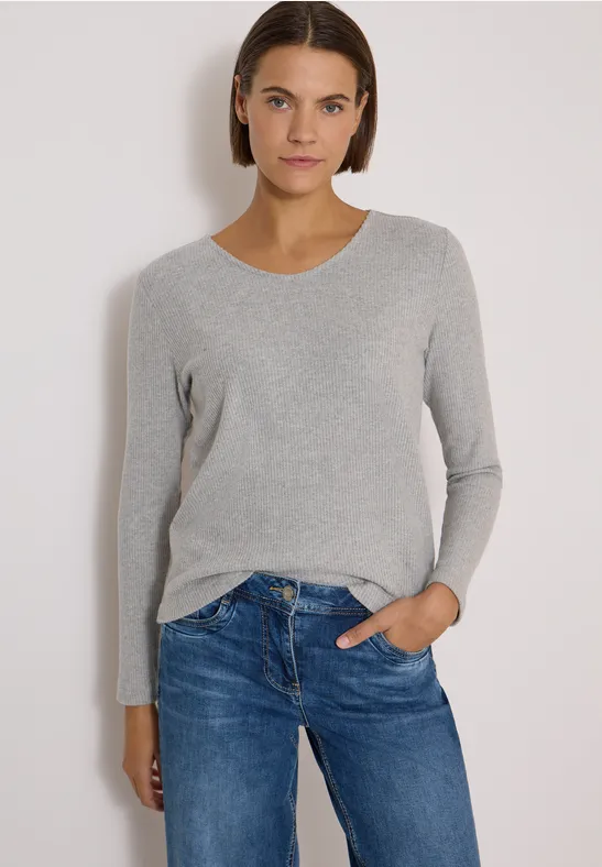 Frau in grauem Pullover und blauen Jeans vor hellem Hintergrund, blickt direkt in die Kamera.