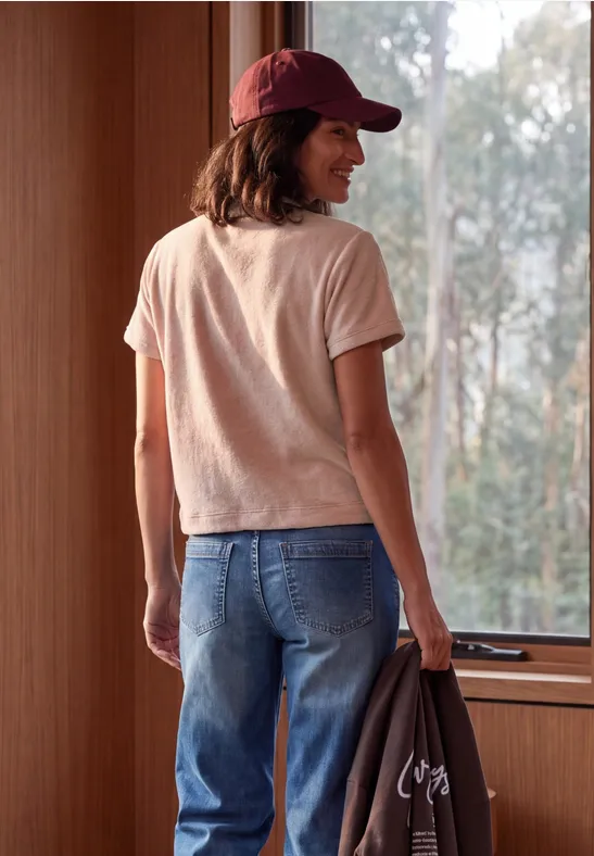 Frau mit roter Kappe, rosa T-Shirt und Jeans hält Jacke und blickt lächelnd aus dem Fenster in Holzhaus.
