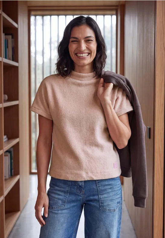Eine lächelnde Frau in Jeans und beigem Pullover steht mit Jacke über der Schulter in einem hellen Flur.