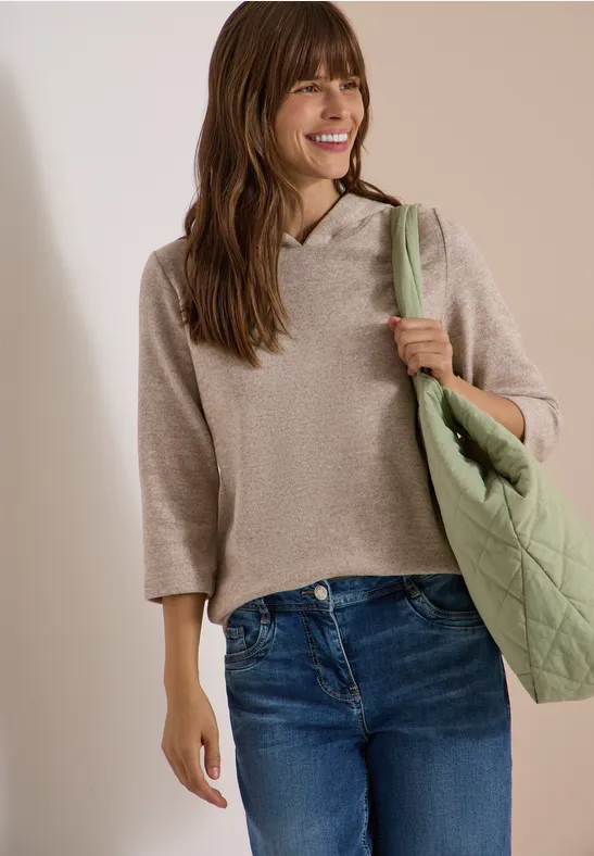 Eine lächelnde Frau trägt beige Kapuzenpullover, blaue Jeans und hält eine grüne Tasche.