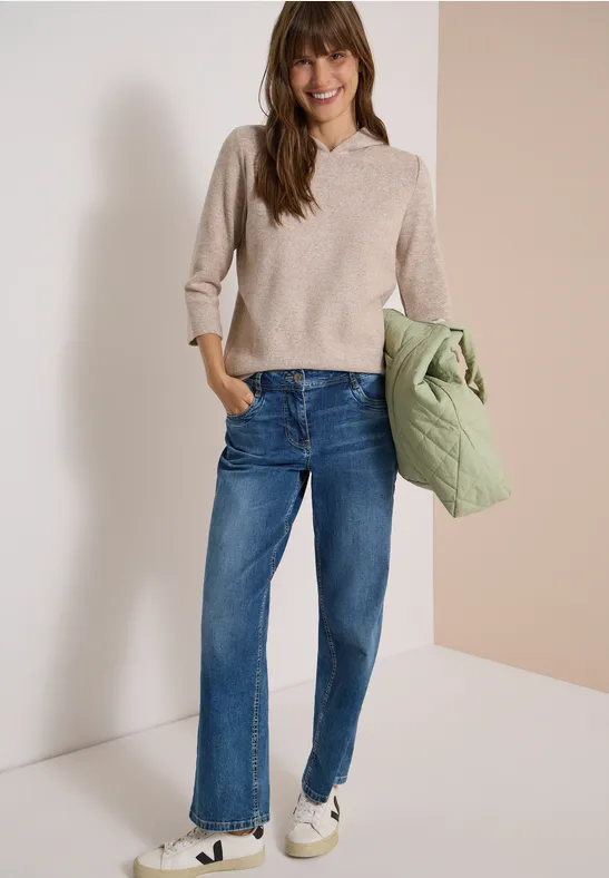 Eine lächelnde Frau trägt einen beigen Pullover, blaue Jeans, weiße Sneaker und hält eine hellgrüne Jacke.