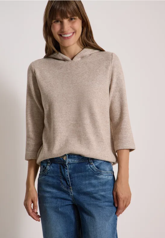 Eine Frau lächelt und trägt einen hellbeigen Kapuzenpullover und blaue Jeans.