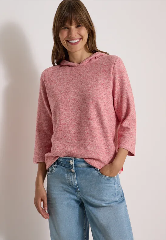 Lächelnde Frau in rosa Kapuzenpullover und blauen Jeans, lässig mit Händen in den Taschen.