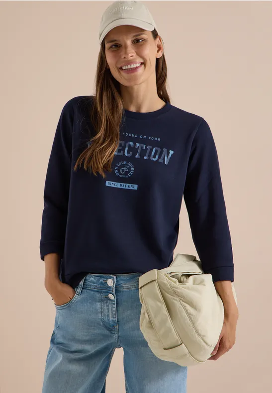 Eine lächelnde Frau in dunkelblauem Sweatshirt mit Schriftzug, Jeans und großer Umhängetasche.