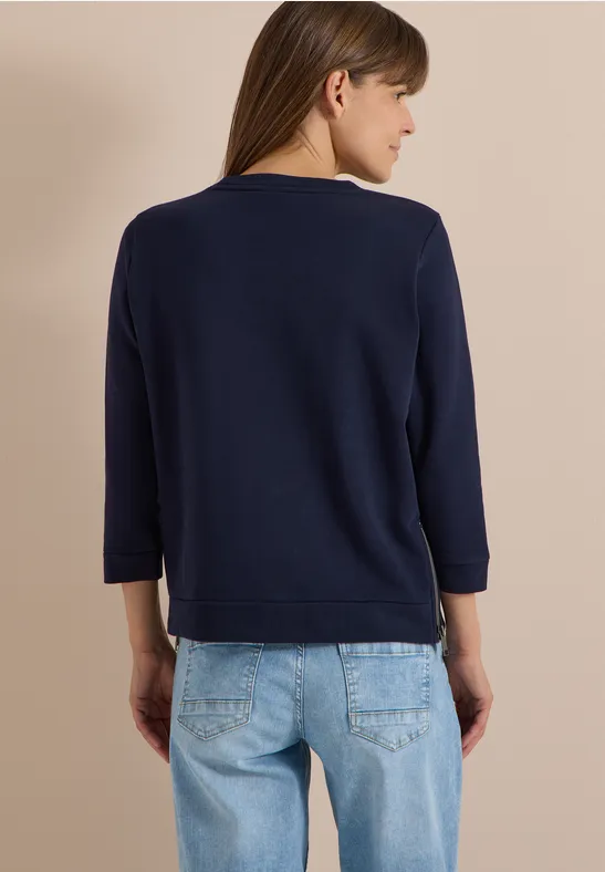 Rückansicht einer Frau im dunkelblauen Langarmshirt und hellblauen Jeans vor beigem Hintergrund.