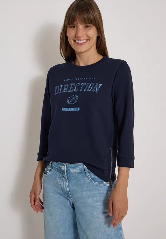 Frau in Jeans und dunkelblauem Shirt mit 'Direction'-Aufdruck, lächelnd vor hellem Hintergrund.