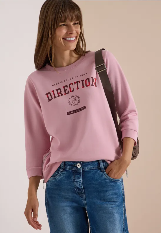 Lächelnde Frau trägt rosa Sweatshirt mit 'DIRECTION'-Schriftzug, blaue Jeans und Schultertasche.