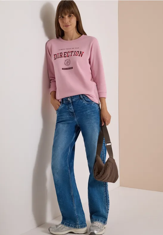 Frau in rosa Sweatshirt mit Aufdruck, blauen Jeans und brauner Schultertasche vor schlichter Kulisse.
