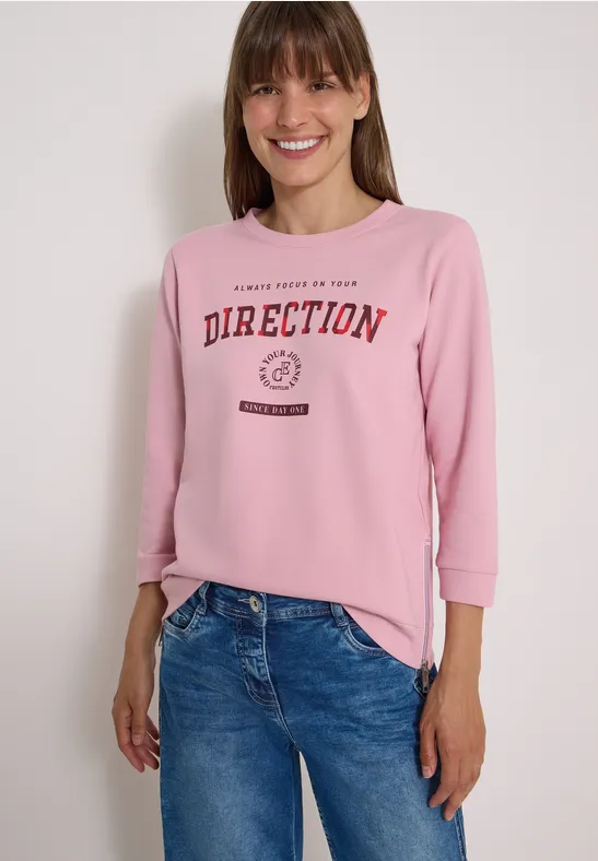 Frau lächelt, trägt ein pinkes Sweatshirt mit „DIRECTION“-Schriftzug und blaue Jeans.