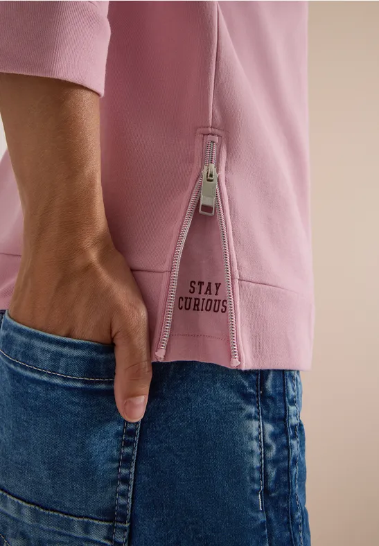 Nahaufnahme eines rosa Oberteils mit seitlichem Reißverschluss, Aufschrift 'STAY CURIOUS' und blauer Jeanshose.