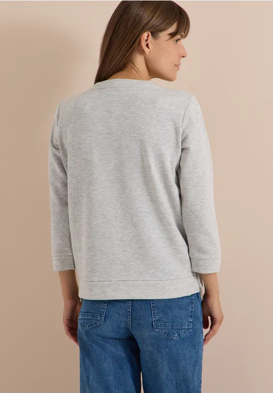 Frau von hinten in grauem Sweatshirt mit 3/4-Ärmeln und blauen Jeans vor hellem Hintergrund.