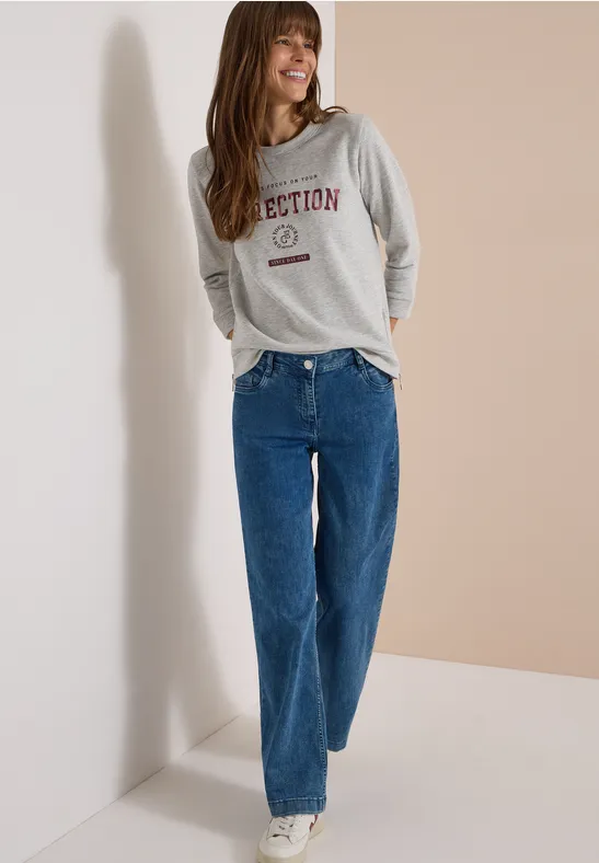 Eine lachende Frau in grauem Sweatshirt und blauen, gerade geschnittenen Jeans vor hellem Hintergrund.