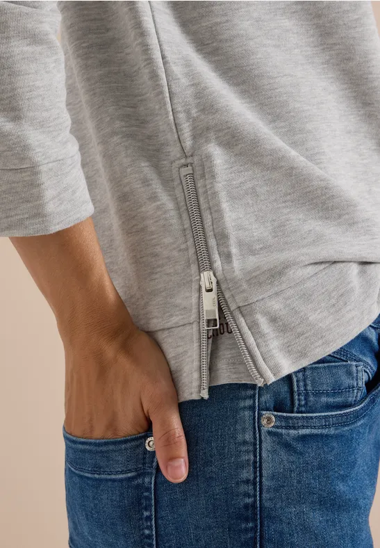 Hand in Jeans-Tasche, darüber graues Oberteil mit seitlichem Reißverschluss.