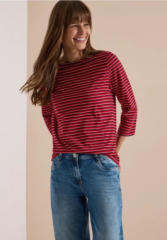Femme souriante en t-shirt rayé rouge et noir et jean bleu, posant devant un fond neutre.