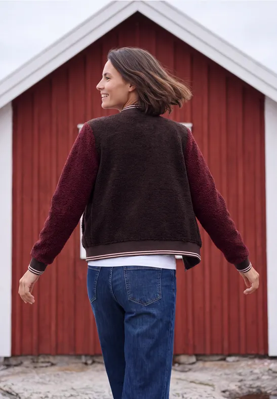 Vrouw met bruin haar in jeans en vest, armen wijd uitgestrekt voor een rode schuur.