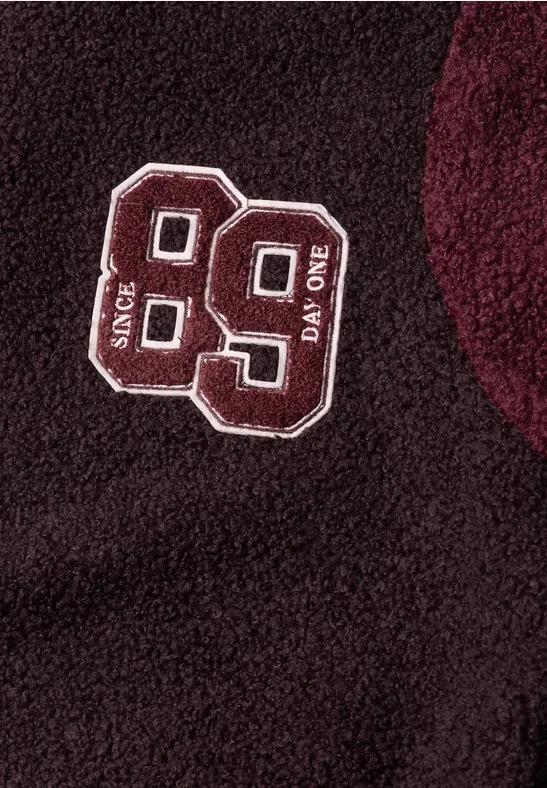 Donkerrood fleece kledingstuk met geborduurde witte '99' patch en verticale tekst 'SINCE' en 'DAY ONE'.