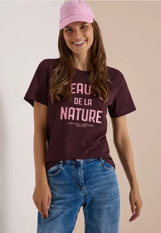 Une femme souriante porte une casquette rose, un t-shirt bordeaux à inscription et un jean bleu.
