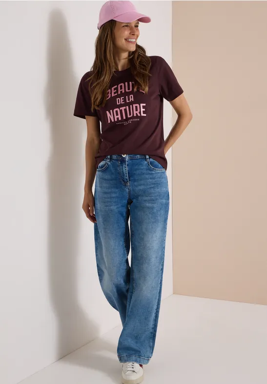 Femme souriante, t-shirt bordeaux imprimé, jean large et casquette rose sur fond clair