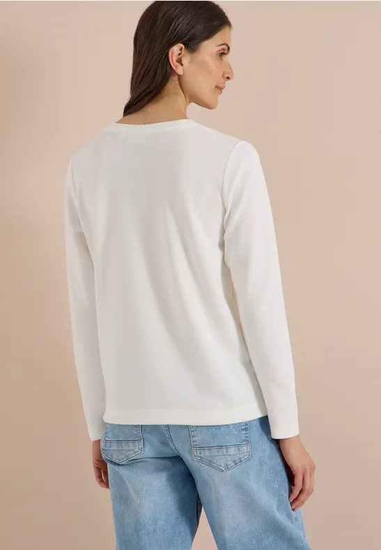 Frau trägt weißes Langarmshirt und blaue Jeans, steht leicht seitlich vor beigem Hintergrund.