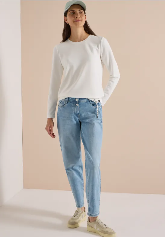 Eine Frau in weißem Langarmshirt, blauen Jeans, Cap und Sneakers steht vor hellem Hintergrund.