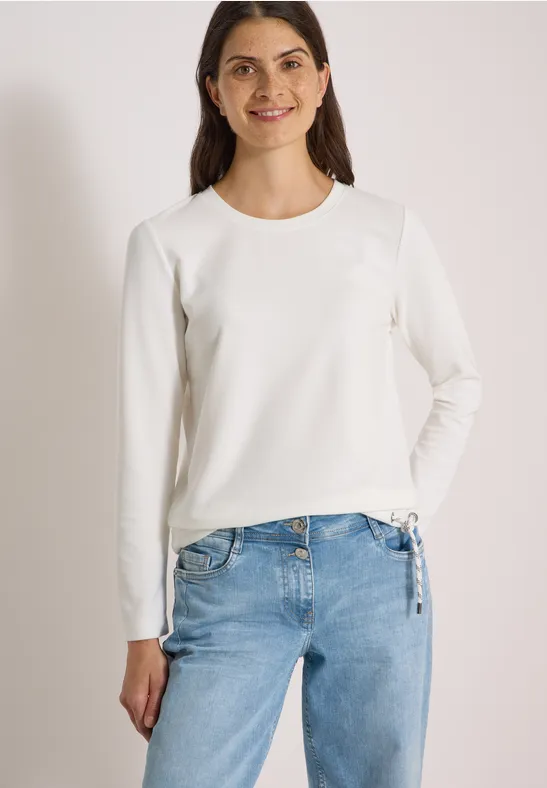 Frau trägt weißen Langarmshirt mit Rundhalsausschnitt und hellblaue Jeans vor neutralem Hintergrund.