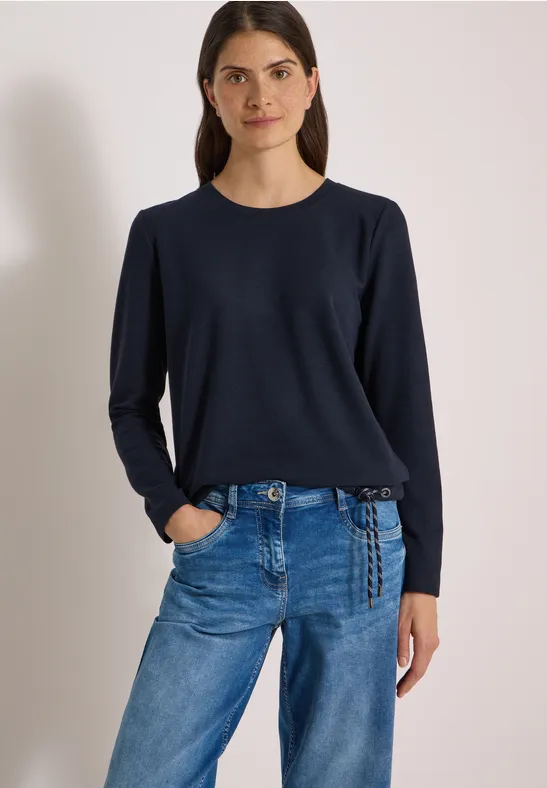 Kobieta w granatowej bluzce z długim rękawem i niebieskich dżinsach na jasnym tle.