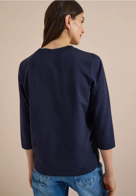Rückenansicht einer Frau in navyblauem 3/4-Arm-Shirt und blauen Jeans vor beigem Hintergrund.