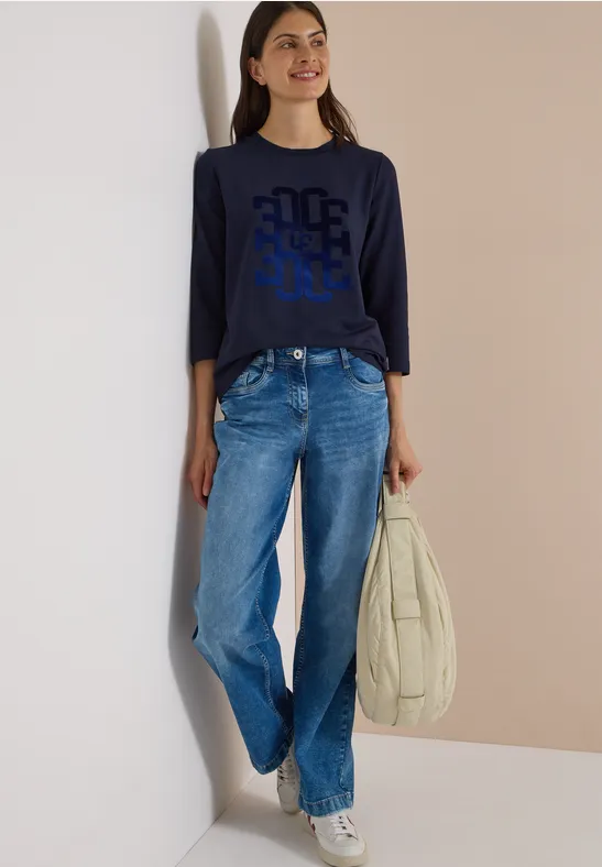 Frau lehnt an Wand, trägt marineblaues Langarmshirt mit Grafik, weite Jeans, weiße Sneaker und große Tasche.