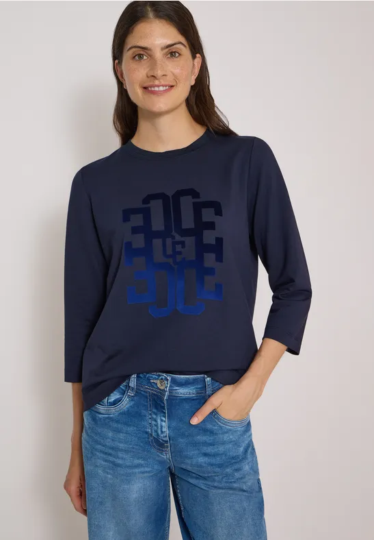 Eine lächelnde Frau in blauem Langarmshirt mit Grafik-Print und Jeans, eine Hand in der Hosentasche.