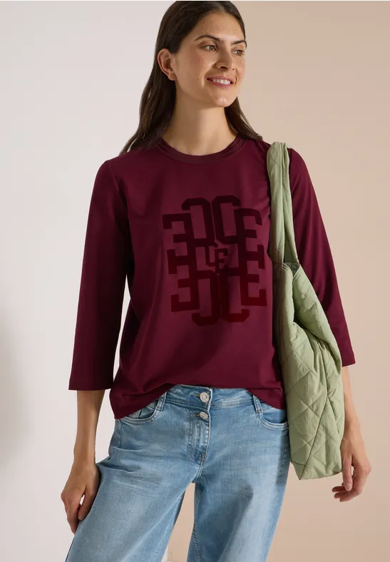 Frau mit burgunderfarbenem Shirt mit 3/4-Ärmeln, Jeans und grüner Stepp-Umhängetasche, lächelnd.